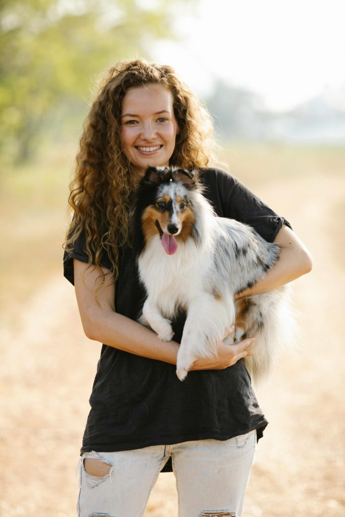 pexels-photo-7210525-7210525 Cheerful woman with curly hair holding a blue merle Shetland sheepdog in a sunny outdoor setting.